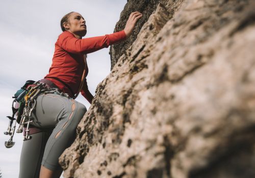 A person is climbing a large rock face, equipped with climbing gear and a harness, focused and reaching upwards.