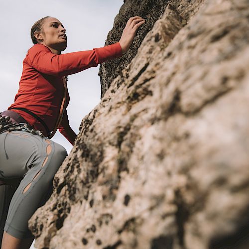 A person is climbing a large rock face, equipped with climbing gear and a harness, focused and reaching upwards.