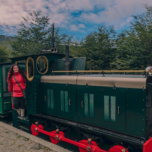 A person in a red jacket stands beside a green and black train in a natural setting with trees and a cloudy sky.