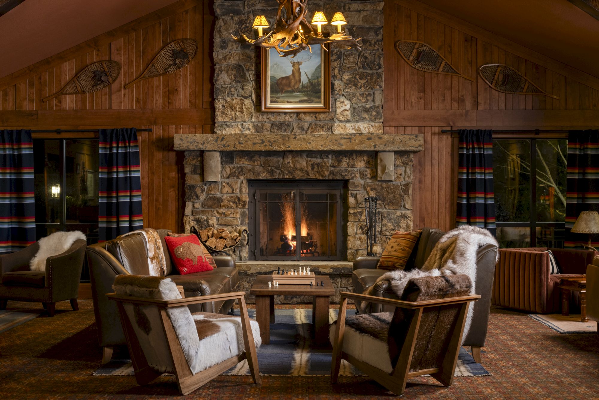A cozy room with a stone fireplace, surrounded by wooden walls, chairs, a chess board, and warm lighting under a framed picture.
