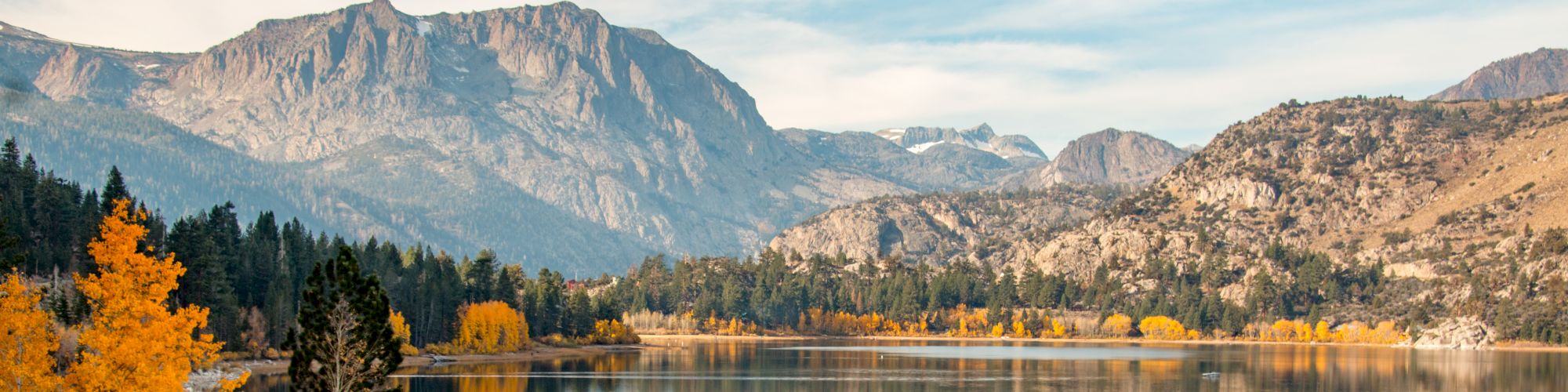 A serene lake reflecting a mountain range, surrounded by vibrant autumn trees under a partly cloudy sky.