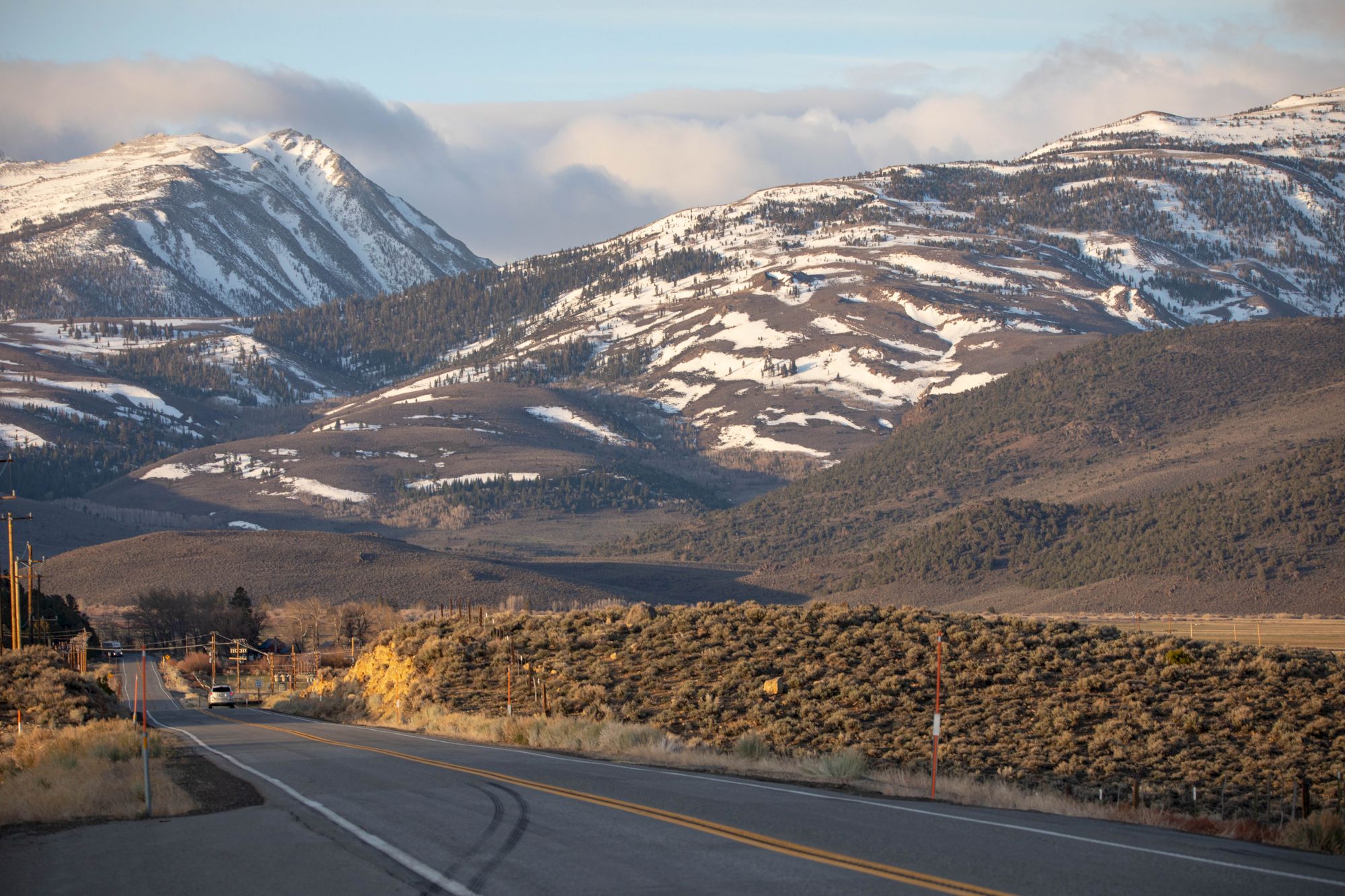 A scenic road leads towards snow-capped mountains under a cloudy sky, surrounded by rugged terrain and sparse vegetation.