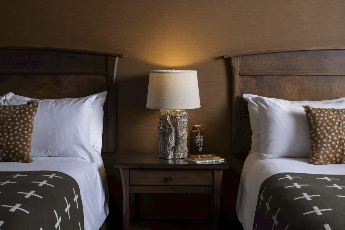 The image shows two beds with wooden headboards, each with pillows and blankets. A lamp on a nightstand stands between the beds.