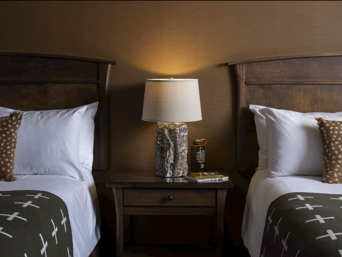 The image shows two beds with wooden headboards, each with pillows and blankets. A lamp on a nightstand stands between the beds.
