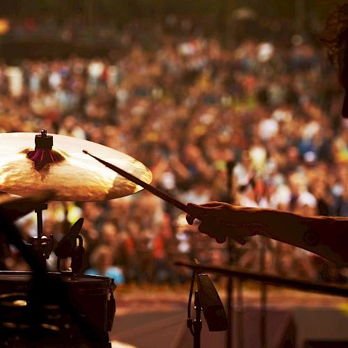 A drummer silhouetted against a large, blurred crowd during a live performance, with cymbals and drumsticks visible in the foreground.