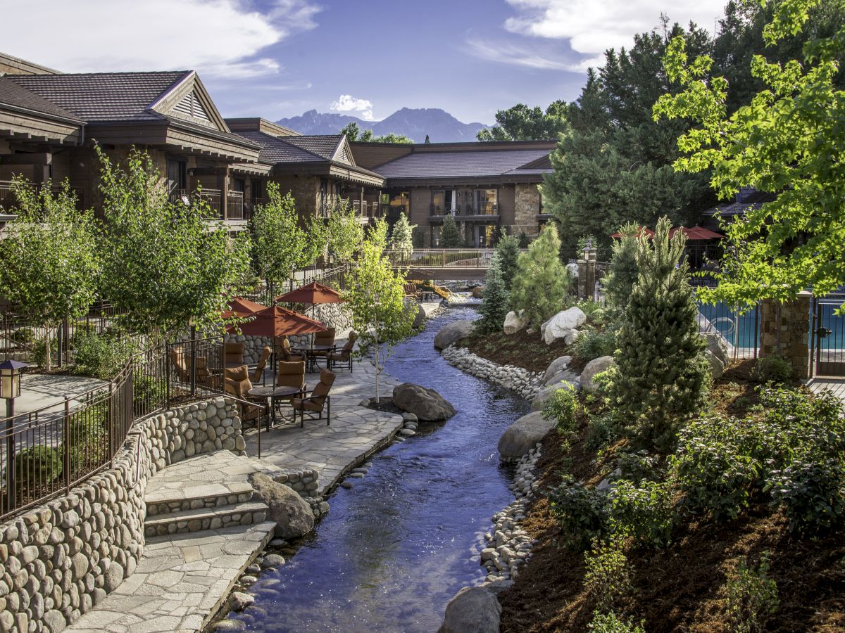 A scenic view of a landscaped area features a flowing stream, patio seating, lush greenery, and a building in the background.