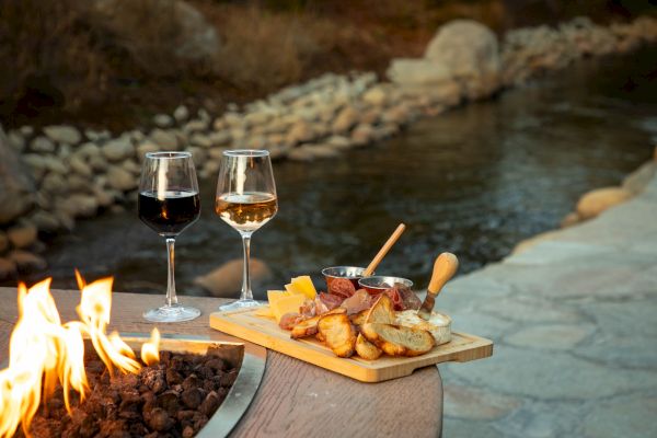 Two glasses of wine and a charcuterie board are set on a table by a fire pit, beside a river with rocks.