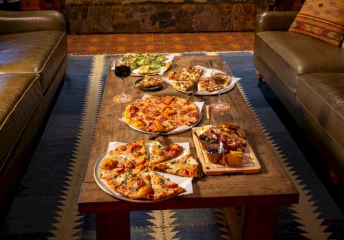 A cozy room with a stone fireplace, sofas, and a wooden table filled with pizzas and snacks, creating a warm and inviting setting.