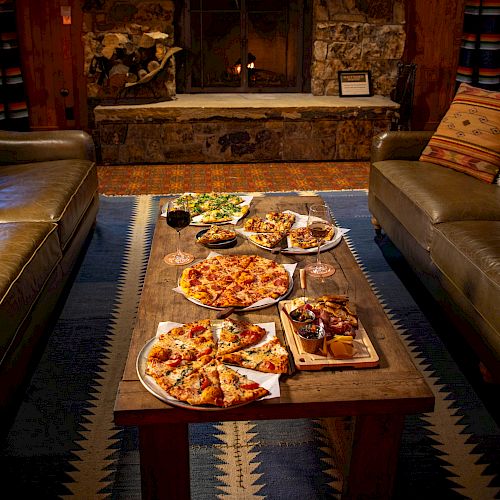 A cozy room with a stone fireplace, sofas, and a wooden table filled with pizzas and snacks, creating a warm and inviting setting.