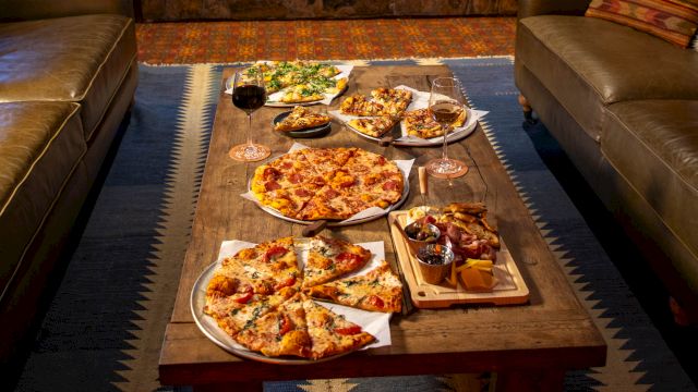 A cozy room with a stone fireplace, sofas, and a wooden table filled with pizzas and snacks, creating a warm and inviting setting.