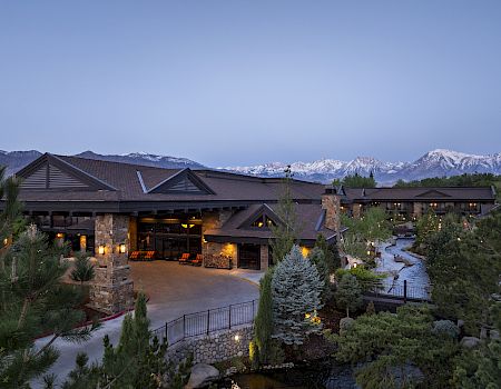 A modern resort lodge with lit patio, landscaped gardens, and a winding stream, set against snowcapped mountains at dusk.