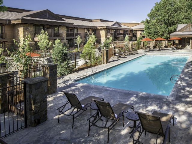 The image shows a sunny outdoor pool area with several loungers on a stone deck, surrounded by trees and a fenced area.