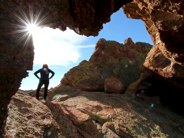 A person stands near a rock formation with a sunburst peeking through, under a clear blue sky.