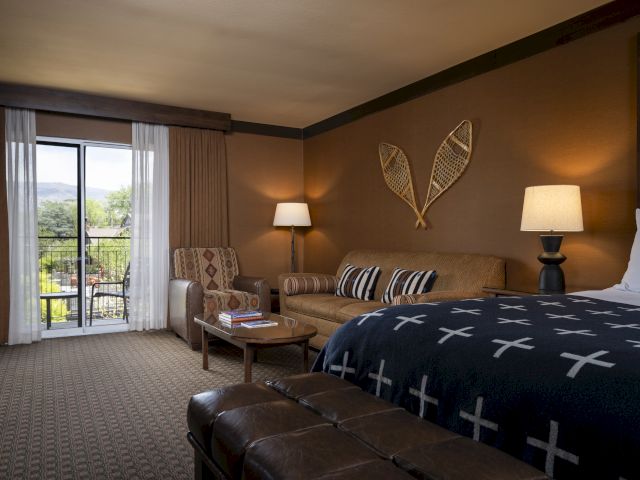 A cozy hotel room with a bed, couch, lamps, and decorative snowshoes on the wall. Sliding doors open to a balcony with a scenic view.