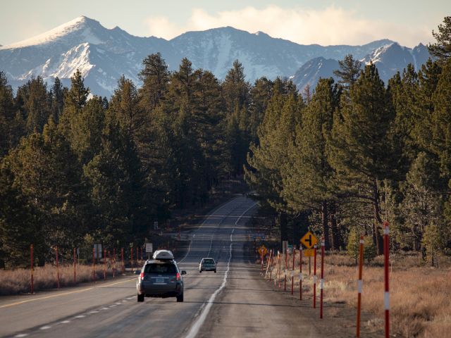A scenic road flanked by trees leads toward snow-capped mountains, with cars driving along and clear skies above.