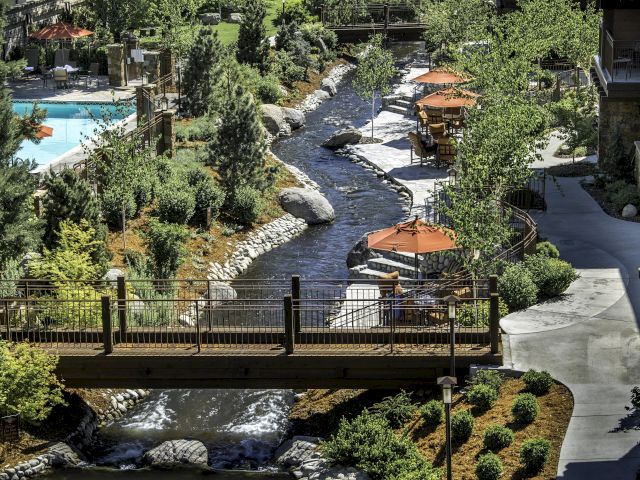The image shows a scenic hotel courtyard with a flowing stream, surrounded by greenery, outdoor seating, and a view of a swimming pool.