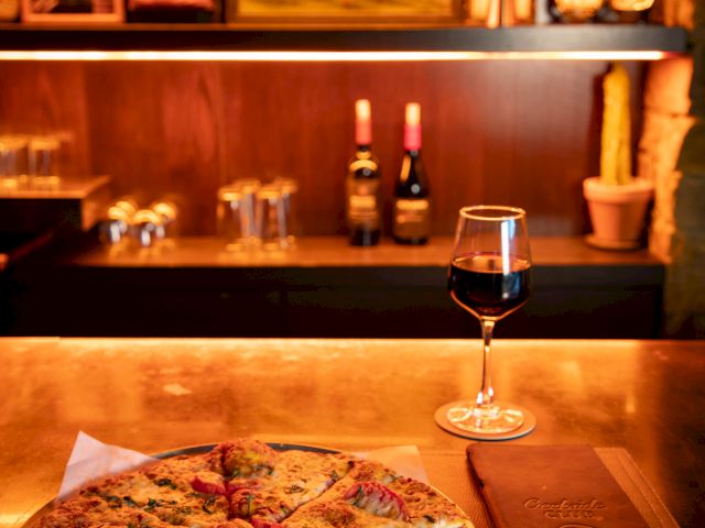 A delicious pizza is on a table with a glass of red wine, menu, and a cozy background featuring framed art and wine bottles.