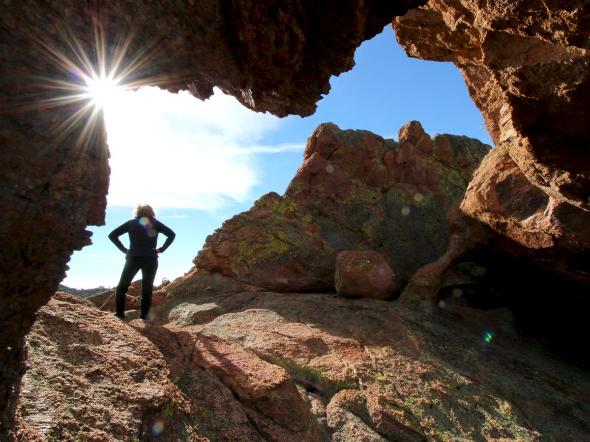 A person stands in a rocky area under a sunny sky, casting a silhouette in the sunlight streaming through rocks.