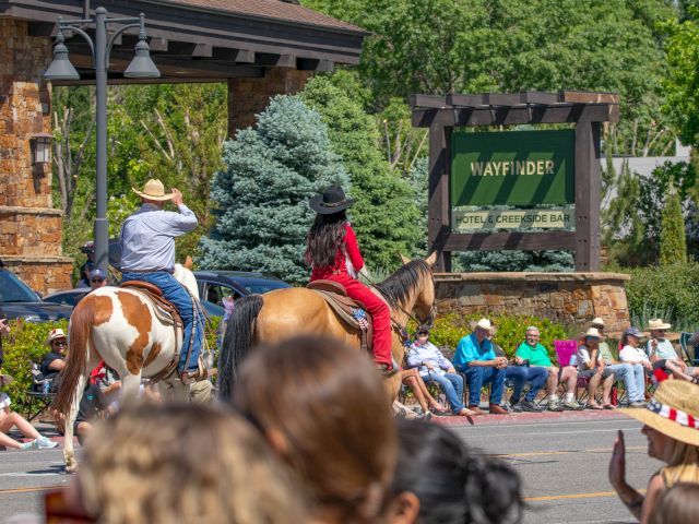 Two people on horseback in a parade; spectators sit along the street near a sign reading "Wayfinder Hotel & Event Center."