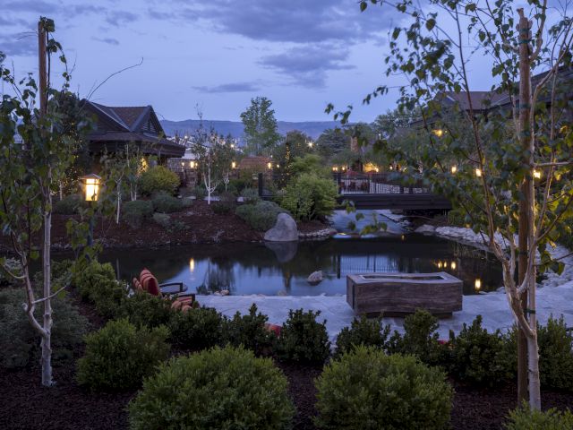 A serene garden scene at dusk features a pond, trees, and a small bridge, with warm lights illuminating the tranquil setting.