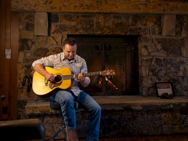A person is playing a guitar while sitting in front of a stone fireplace with a painting above it.