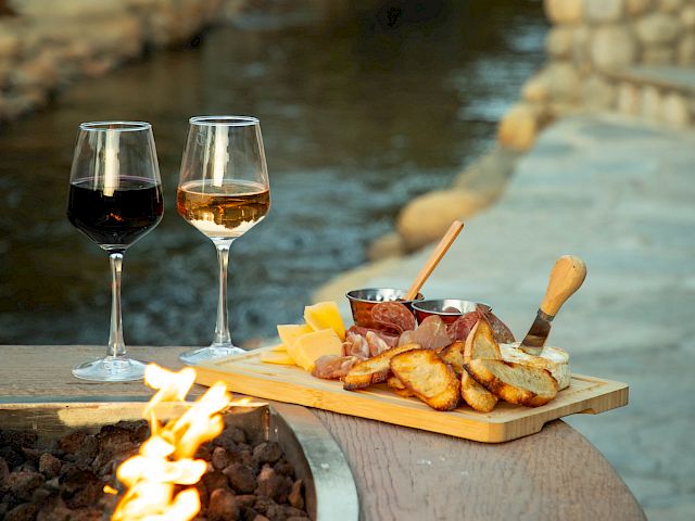 Two glasses of wine and a charcuterie board by a fire pit next to a serene creek setting.