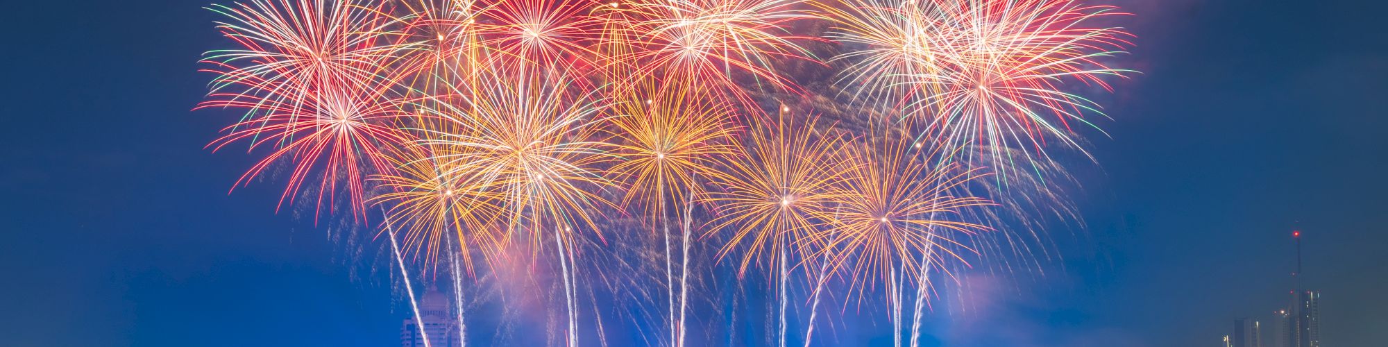 A vibrant fireworks display lights up the night sky above a river, with boats visible on the water and a bridge in the background.