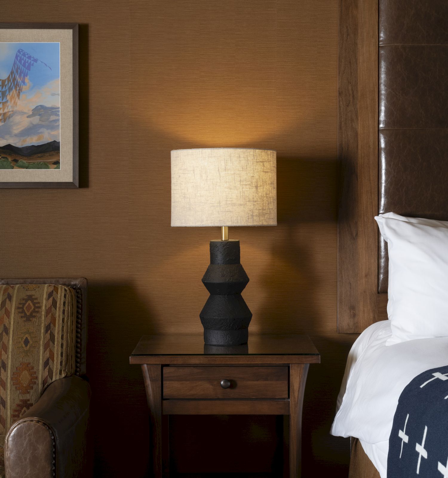 A cozy room with a patterned armchair, decorative lamp on a wooden table, framed artwork, and a bed with a cross-patterned blanket.