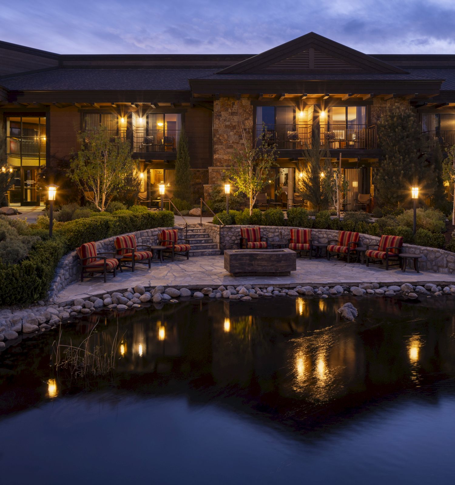 The image shows a large, illuminated house at dusk, with a patio, seating area, and a pond reflecting the lights in front.