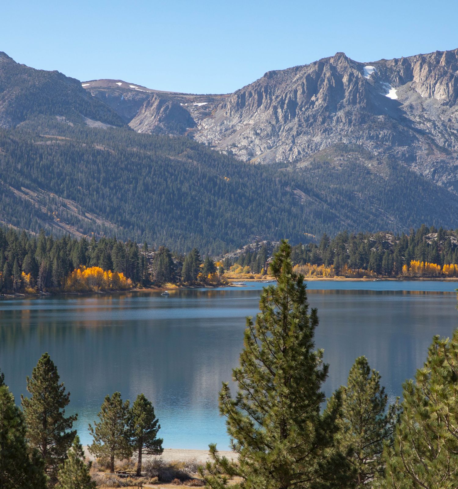 A serene lake surrounded by pine trees and mountains, with a hint of autumn colors in the distance under a clear sky.