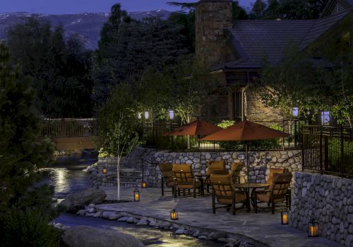 A peaceful outdoor patio with tables, chairs, and umbrellas by a stream, surrounded by trees and lanterns, set in evening light.