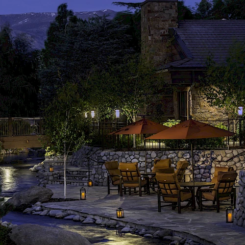 A peaceful outdoor patio with tables, chairs, and umbrellas by a stream, surrounded by trees and lanterns, set in evening light.