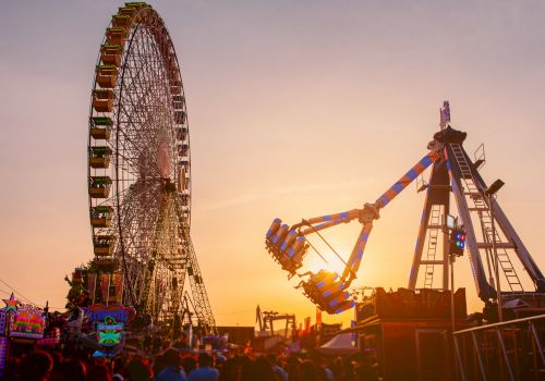 The image shows a Ferris wheel and a swinging amusement ride against a sunset backdrop with a crowd of people below.