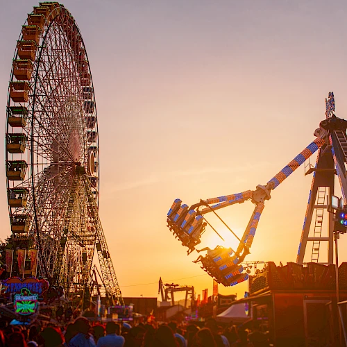 The image shows a Ferris wheel and a swinging amusement ride against a sunset backdrop with a crowd of people below.