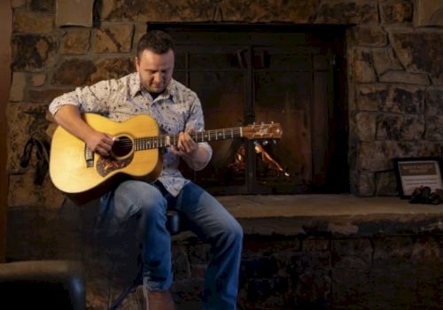 A man in a checked shirt and jeans sits by a stone fireplace, playing an acoustic guitar.