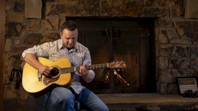 A man in a checked shirt and jeans sits by a stone fireplace, playing an acoustic guitar.