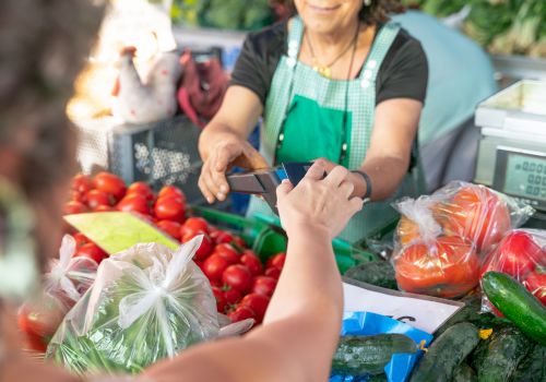 A shopper pays a vendor with a card while a vendor counts money, surrounded by fresh produce like tomatoes, peppers, and greens.