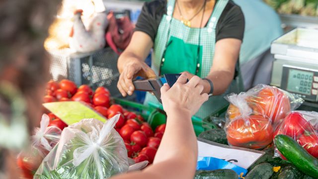 A shopper pays a vendor with a card while a vendor counts money, surrounded by fresh produce like tomatoes, peppers, and greens.
