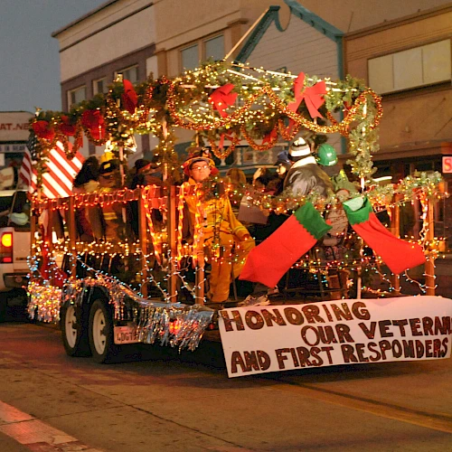 A decorated parade float with glittering lights and a banner honoring veterans and first responders, driving through a small town street at dusk.
