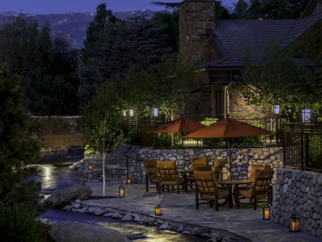 A cozy outdoor patio with tables, chairs, and umbrellas surrounded by trees and lanterns, set against a stone building and twilight sky.