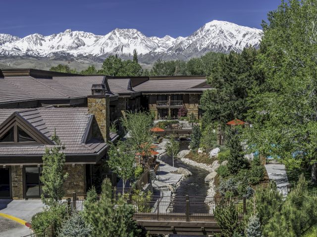 The image shows a building surrounded by greenery, with snow-capped mountains in the background under a clear blue sky.