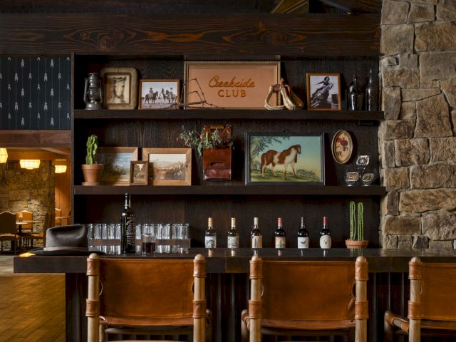 The image shows a rustic bar area with wooden chairs, framed pictures, bottles, and stone wall decor under a sloped ceiling.