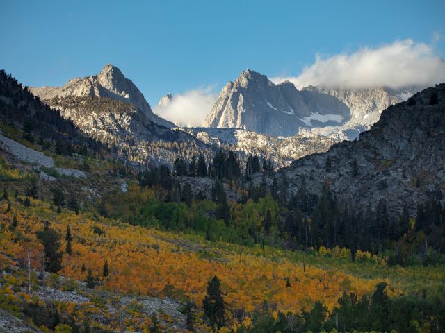 A scenic mountain landscape with snow-capped peaks, forests, and colorful autumn foliage under a blue sky with scattered clouds.
