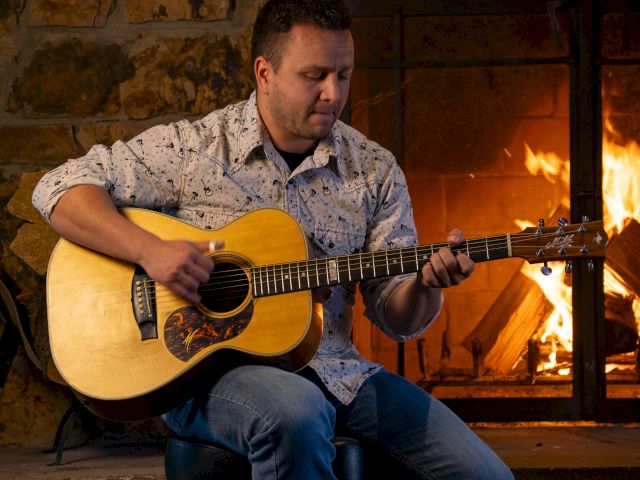 A person is playing an acoustic guitar in front of a lit fireplace, sitting in a cozy, rustic setting.