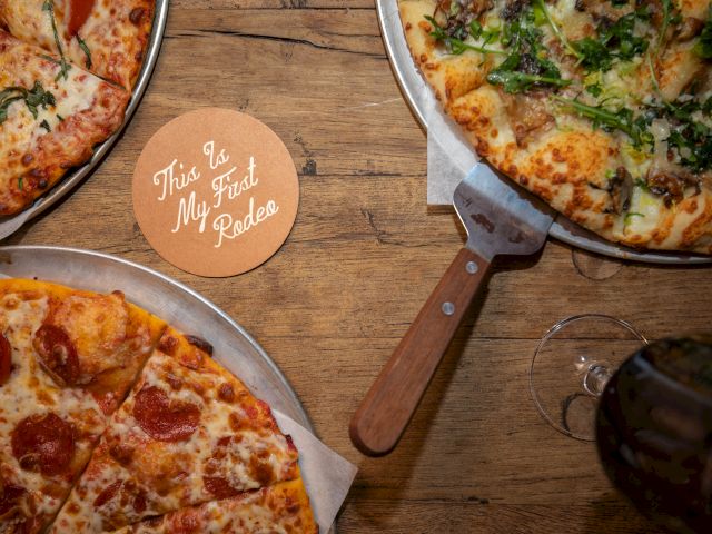 Three pizzas on pans and a coaster with the text "This is My 3rd Pizza" are on a wooden table.