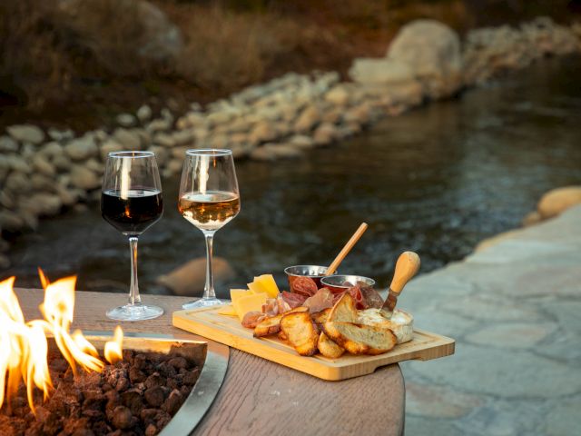 A cozy outdoor scene with two wine glasses and a cheese board by a fire pit, overlooking a serene creek setting.