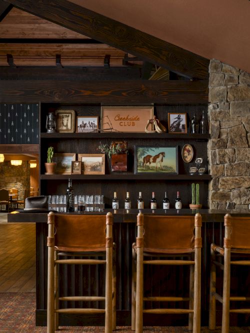 Cozy bar area with wooden stools, leather accents, stone wall, and assorted vintage decor. Bottles line the counter beneath framed pictures.