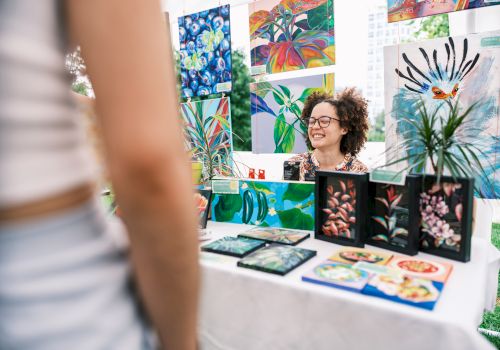 A woman vendor sits at a table with colorful botanical art and prints on display, greeting a customer whose arm is visible in the foreground.