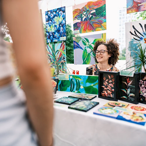 A woman vendor sits at a table with colorful botanical art and prints on display, greeting a customer whose arm is visible in the foreground.