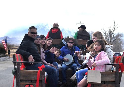 A group of people ride a wooden wagon, with musicians in front, a child in pink, and small American flags in the background, festive and casual.